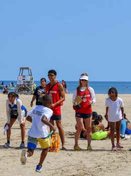 Junior Guard participants running a relay race on a sandy beach, supervised by lifeguards.