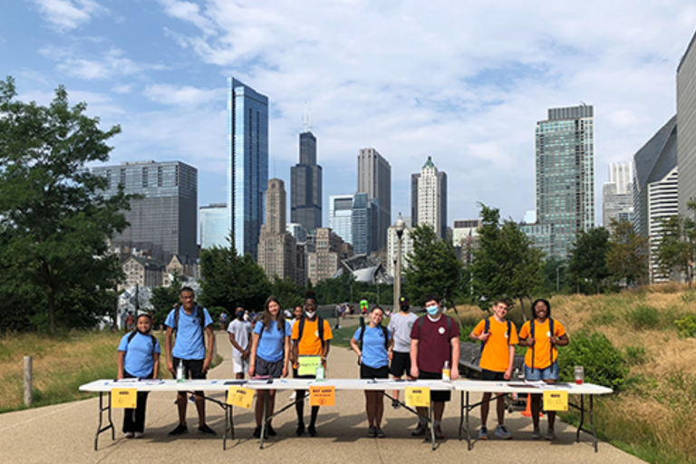 Students stand at a table with the Chicago skyline in the background.
