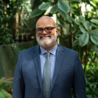 Smiling man in a blue suit and gray tie poses in front of greenery.