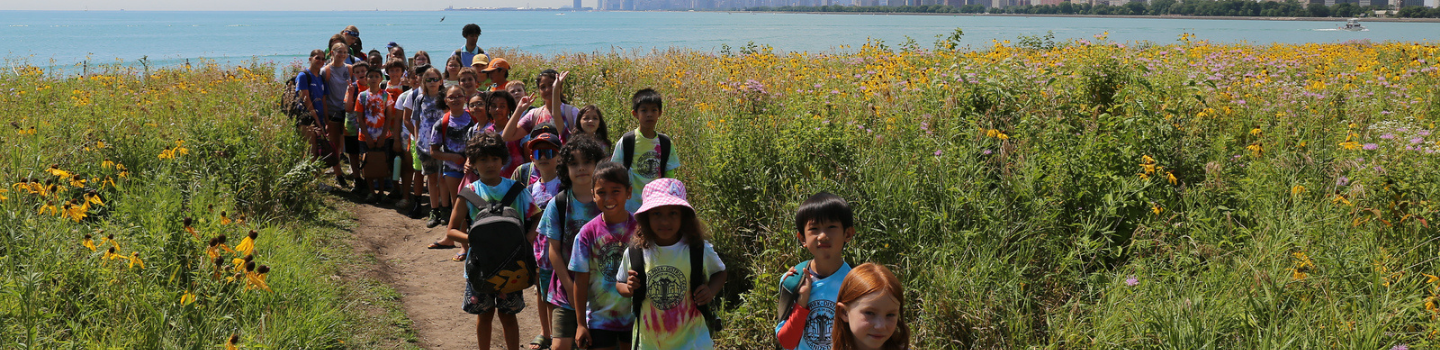 Children hike on a trail through wildflowers with a city skyline across the water.