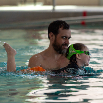 Dad helps child with orange floatie learn to swim.