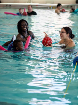 Two women and a toddler playing with a red ball during a swim lesson.