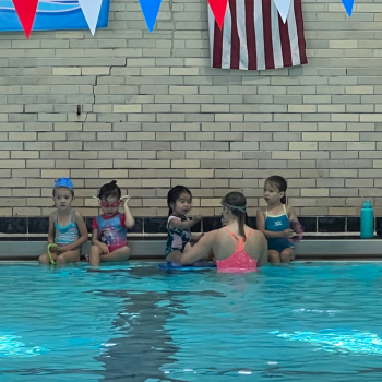 Swim instructor and four young children sit at the edge of a pool.