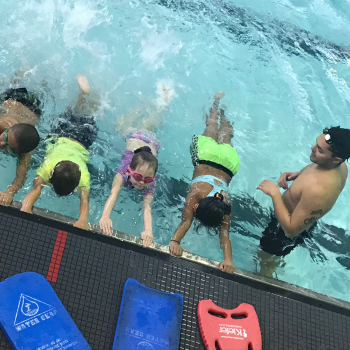 Children hold pool edge during swim lesson with instructor.