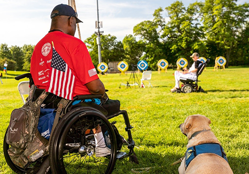 Two wheelchair users at an archery range, one with a service dog.