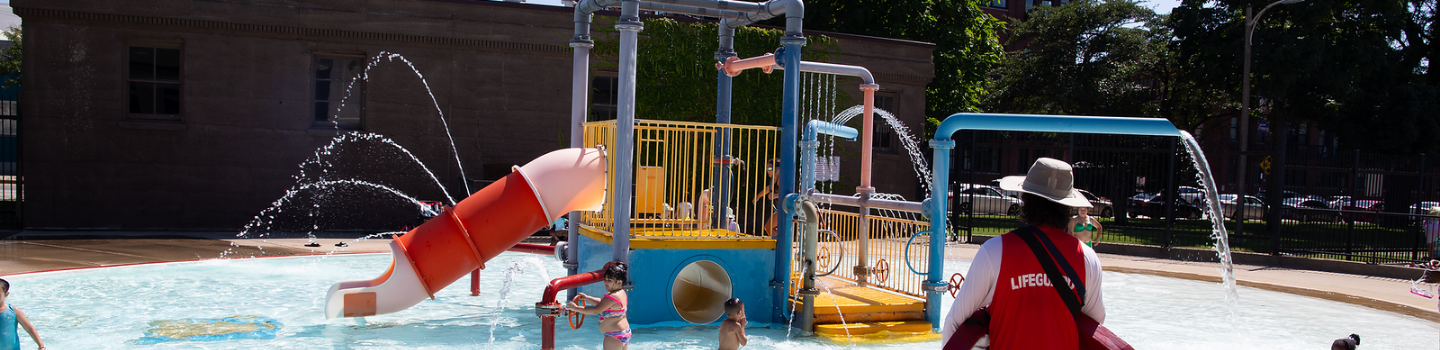 Children play on a colorful splash pad; a lifeguard watches.